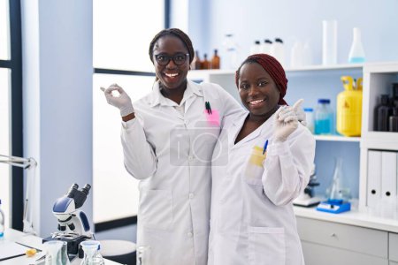 Téléchargez les photos : Deux femmes africaines travaillant au laboratoire scientifique souriant heureux pointant avec la main et le doigt sur le côté - en image libre de droit