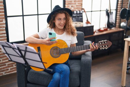 Téléchargez les photos : Jeune belle musicienne hispanique jouant de la guitare classique en utilisant un smartphone au studio de musique - en image libre de droit