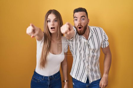 Photo for Young couple standing over yellow background pointing with finger surprised ahead, open mouth amazed expression, something on the front - Royalty Free Image