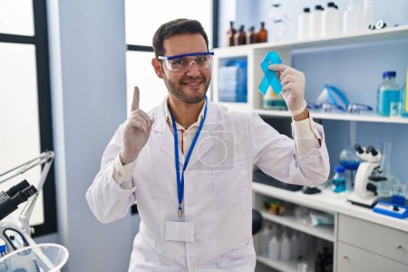 Téléchargez les photos : Jeune homme hispanique avec barbe travaillant au laboratoire scientifique tenant un ruban bleu surpris par une idée ou une question pointant du doigt avec un visage heureux, numéro un - en image libre de droit