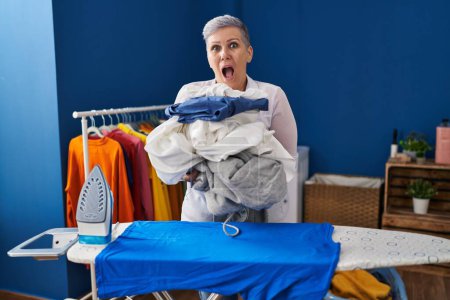 Foto de Mujer de mediana edad planchando ropa en casa asustada y sorprendida con expresión sorpresa y asombro, miedo y cara excitada. - Imagen libre de derechos