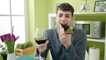 Téléchargez les photos : Jeune homme hispanique boire un verre de vin envoyer un message vocal avec smartphone à la salle à manger - en image libre de droit