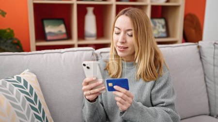 Foto de Mujer rubia joven de compras con teléfono inteligente y tarjeta de crédito sentado en el sofá en casa - Imagen libre de derechos