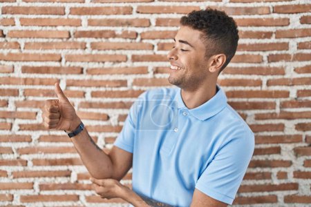 Téléchargez les photos : Jeune homme brésilien debout au-dessus du mur de briques regardant fier, souriant faire les pouces geste vers le haut sur le côté - en image libre de droit
