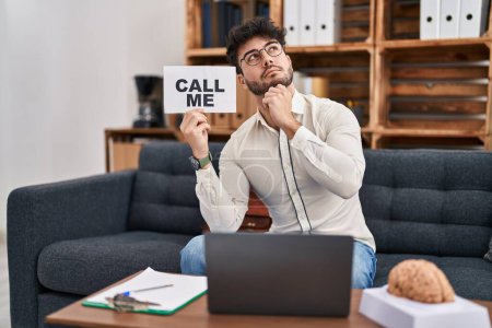Photo for Hispanic man with beard working at therapy office holding call me sign serious face thinking about question with hand on chin, thoughtful about confusing idea - Royalty Free Image