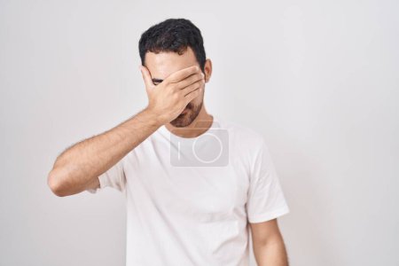Photo for Handsome hispanic man standing over white background covering eyes with hand, looking serious and sad. sightless, hiding and rejection concept - Royalty Free Image