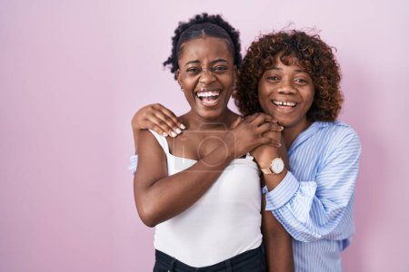 Téléchargez les photos : Deux femmes africaines debout sur fond rose souriant et riant fort à haute voix parce que drôle blague folle. - en image libre de droit