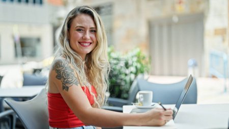 Téléchargez les photos : Jeune femme blonde utilisant un ordinateur portable prenant des notes souriantes à la terrasse du café - en image libre de droit
