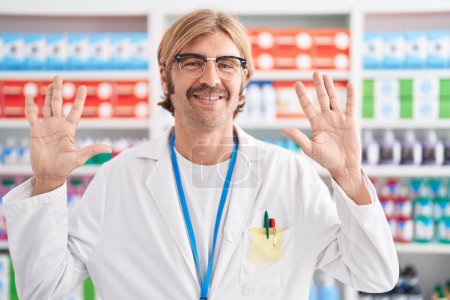 Téléchargez les photos : Homme caucasien avec moustache travaillant à la pharmacie pharmacie montrant et pointant vers le haut avec les doigts numéro dix tout en souriant confiant et heureux. - en image libre de droit