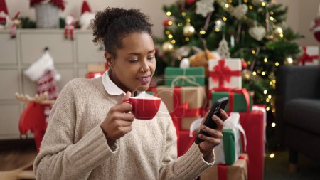 Photo for African american woman using smartphone drinking coffee sitting by christmas tree at home - Royalty Free Image