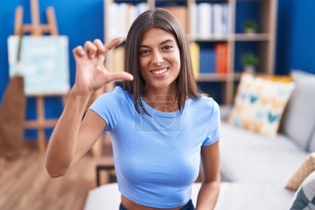 Photo for Brunette young woman sitting on the sofa at home smiling and confident gesturing with hand doing small size sign with fingers looking and the camera. measure concept. - Royalty Free Image