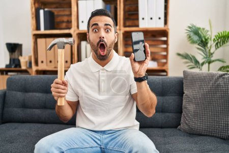 Photo for Young hispanic man with beard holding hammer and broken smartphone showing cracked screen afraid and shocked with surprise and amazed expression, fear and excited face. - Royalty Free Image