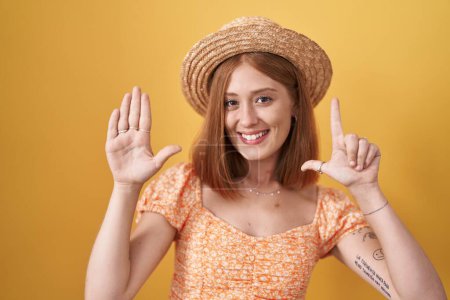 Téléchargez les photos : Jeune rousse femme debout sur fond jaune portant chapeau d'été montrant et pointant vers le haut avec les doigts numéro sept tout en souriant confiant et heureux. - en image libre de droit