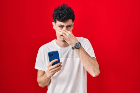 Photo for Hispanic man using smartphone over red background smelling something stinky and disgusting, intolerable smell, holding breath with fingers on nose. bad smell - Royalty Free Image