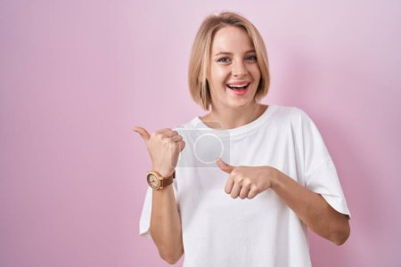 Photo for Young caucasian woman standing over pink background pointing to the back behind with hand and thumbs up, smiling confident - Royalty Free Image
