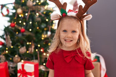 Téléchargez les photos : Adorable fille blonde souriant confiant debout par arbre de Noël à la maison - en image libre de droit
