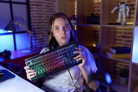 Foto de Joven mujer caucásica sosteniendo teclado gamer expresión despistada y confusa. concepto de duda. - Imagen libre de derechos