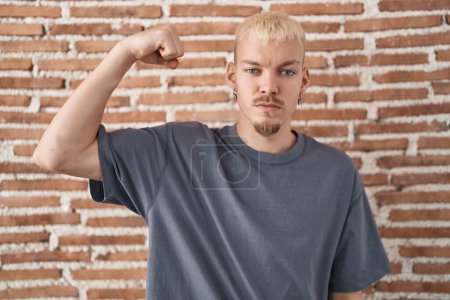Foto de Joven caucásico hombre de pie sobre ladrillos pared fuerte persona mostrando brazo músculo, confiado y orgulloso de poder - Imagen libre de derechos
