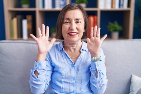 Foto de Mujer hispana de mediana edad sentada en el sofá en casa mostrando y señalando con los dedos el número nueve mientras sonríe confiada y feliz. - Imagen libre de derechos