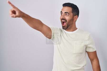 Photo for Hispanic man with beard standing over isolated background pointing with finger surprised ahead, open mouth amazed expression, something on the front - Royalty Free Image