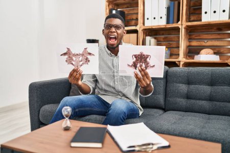 Photo for African american man working with rorschach test at psychology clinic angry and mad screaming frustrated and furious, shouting with anger. rage and aggressive concept. - Royalty Free Image