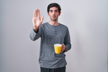 Photo for Young hispanic man wearing pajama drinking a cup of coffee doing stop sing with palm of the hand. warning expression with negative and serious gesture on the face. - Royalty Free Image