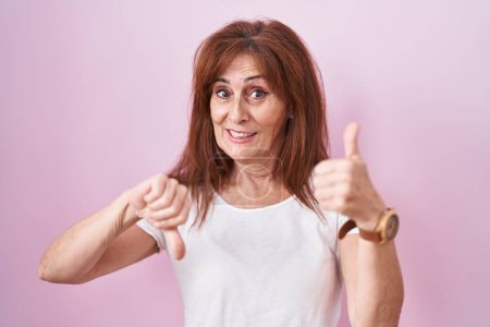 Foto de Middle age woman standing over pink background doing thumbs up and down, disagreement and agreement expression. crazy conflict - Imagen libre de derechos