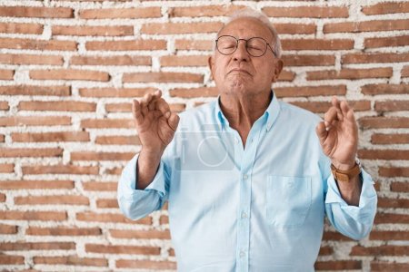 Téléchargez les photos : Homme âgé avec les cheveux gris debout sur le mur de briques détendu et souriant avec les yeux fermés faisant geste de méditation avec les doigts. concept de yoga. - en image libre de droit