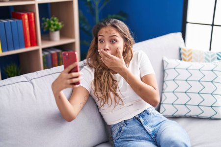 Téléchargez les photos : Jeune femme hispanique faisant appel vidéo avec smartphone couvrant la bouche avec la main, choquée et effrayée par l'erreur. expression surprise - en image libre de droit