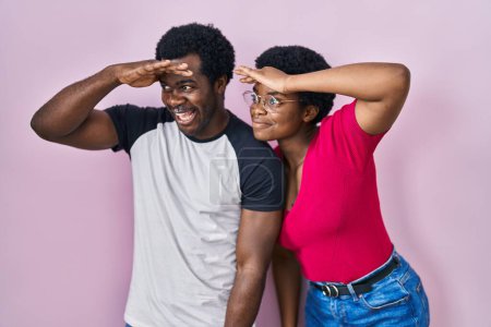 Foto de Joven pareja afroamericana de pie sobre fondo rosa muy feliz y sonriente mirando lejos con la mano sobre la cabeza. concepto de búsqueda. - Imagen libre de derechos