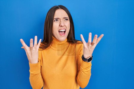 Photo for Young brunette woman standing over blue background crazy and mad shouting and yelling with aggressive expression and arms raised. frustration concept. - Royalty Free Image