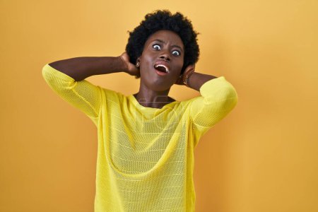 Foto de Mujer joven africana de pie sobre el estudio amarillo loco y asustado con las manos en la cabeza, miedo y sorprendido de choque con la boca abierta - Imagen libre de derechos