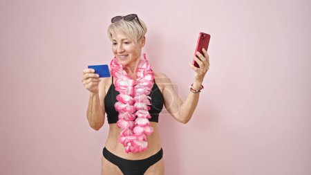 Téléchargez les photos : Femme blonde du moyen âge portant un bikini et hawaiian lei shopping avec smartphone et carte de crédit sur fond rose isolé - en image libre de droit
