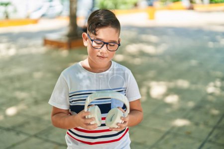 Téléchargez les photos : Adorable garçon hispanique tenant des écouteurs avec une expression détendue au parc - en image libre de droit