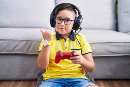 Foto de Joven niño hispano jugando videojuego sosteniendo controlador usando auriculares apuntando hacia atrás con la mano y los pulgares hacia arriba, sonriendo confiado - Imagen libre de derechos