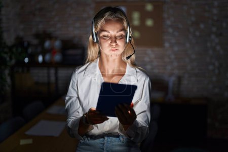 Photo for Young blonde woman working at the office at night puffing cheeks with funny face. mouth inflated with air, crazy expression. - Royalty Free Image