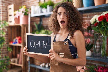 Photo for Hispanic woman with curly hair working at florist holding open sign afraid and shocked with surprise and amazed expression, fear and excited face. - Royalty Free Image
