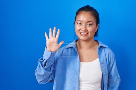 Foto de Asiática joven mujer de pie sobre fondo azul mostrando y señalando hacia arriba con los dedos número cinco mientras sonríe confiado y feliz. - Imagen libre de derechos