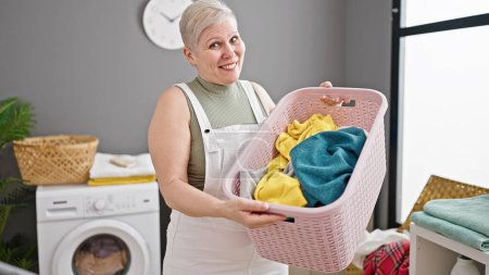 Foto de Mujer de pelo gris de mediana edad sonriendo confiado celebración cesta con ropa en la lavandería - Imagen libre de derechos