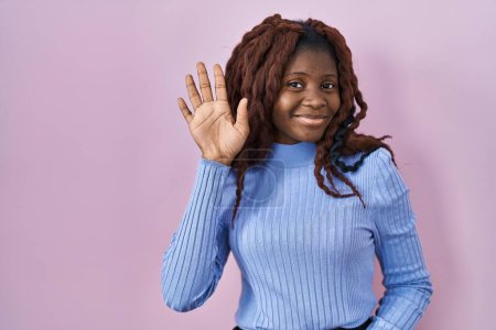 Téléchargez les photos : Femme africaine debout sur fond rose renonçant à dire bonjour heureux et souriant, geste de bienvenue amical - en image libre de droit