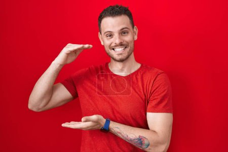 Téléchargez les photos : Jeune homme hispanique debout sur fond rouge geste avec les mains montrant grand et grand signe de taille, symbole de mesure. souriant en regardant la caméra. concept de mesure. - en image libre de droit
