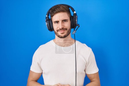 Foto de Hombre hispano con barba escuchando música usando auriculares con las manos juntas y dedos cruzados sonriendo relajado y alegre. éxito y optimismo - Imagen libre de derechos