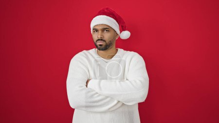 Foto de Hombre afroamericano de pie con los brazos cruzados gesto usando sombrero de Navidad sobre fondo rojo aislado - Imagen libre de derechos