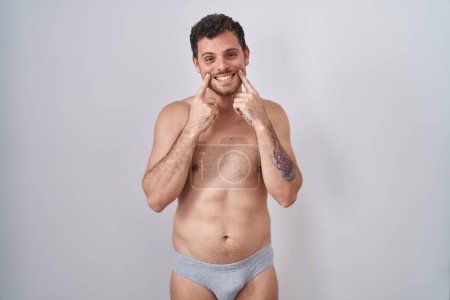 Téléchargez les photos : Jeune homme hispanique debout torse nu portant des sous-vêtements souriant avec la bouche ouverte, les doigts pointant et forçant sourire joyeux - en image libre de droit