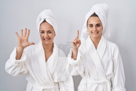 Téléchargez les photos : Femme d'âge moyen et fille portant un peignoir blanc et une serviette montrant et pointant vers le haut avec les doigts numéro six tout en souriant confiant et heureux. - en image libre de droit