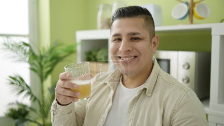 Téléchargez les photos : Jeune homme hispanique souriant confiant tenant verre de jus d'orange à la salle à manger - en image libre de droit