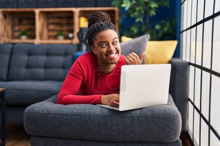 Photo for Young african american with braids working using computer laptop pointing thumb up to the side smiling happy with open mouth - Royalty Free Image
