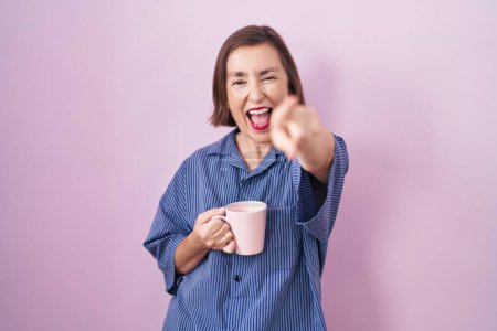 Foto de Mujer hispana de mediana edad bebiendo una taza de café señalándote a ti y a la cámara con los dedos, sonriendo positiva y alegre - Imagen libre de derechos