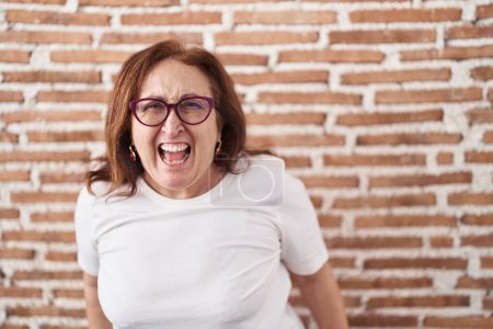 Photo for Senior woman with glasses standing over bricks wall angry and mad screaming frustrated and furious, shouting with anger. rage and aggressive concept. - Royalty Free Image
