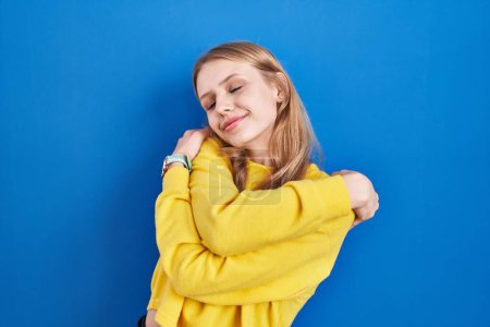 Photo for Young caucasian woman standing over blue background hugging oneself happy and positive, smiling confident. self love and self care - Royalty Free Image